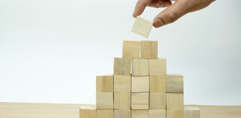 imagine-buddy-FDaCU3etvAc-unsplash Je ziet een houten tafelblad met een witte achtergrond. Op het tafelblad zet een hand houten blokjes neer, in de vorm van een pyramide. Dit symboliseert risicospreiding bij beleggen, omdat je meerdere bouwstenen gebruikt voor het bouwen van je portfolio, en daarmee je risico spreidt.