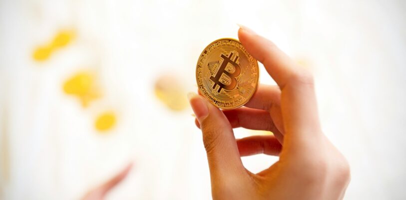 kanchanara-5FLR5KAucEU-unsplash Je ziet een vrij witte achtergrond met op de voorgrond een vrouwelijke hand, die een bitcoin munt vasthoudt.
